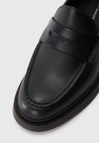 Schwarze Lederloafers verfügen über ein erhöhtes Riemendetail, eine Naht entlang der Kanten und eine flache Sohle mit einer dezenten Textur.