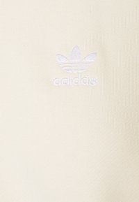 Logotipo blanco de Adidas trefoil bordado sobre un fondo de tela beige claro.