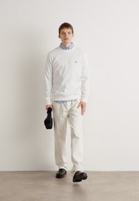 Suéter blanco con logo, sobre una camisa de rayas azul y blanca; emparejado con pantalones blancos y zapatos negros. Sujetando un bolso negro.