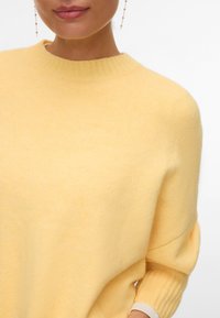 Femme portant un pull côtelé jaune doux à manches longues et des boucles d'oreilles chaînes pendantes, fond blanc uni.