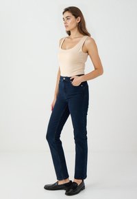 Donna con lunghi capelli castani, che indossa un top senza maniche beige, jeans a vita alta blu scuro e mocassini neri, in piedi su uno sfondo bianco.