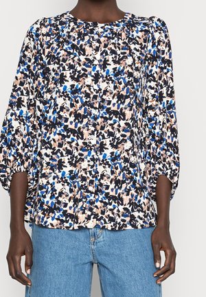 Femme portant un chemisier boutonné à imprimé floral abstrait noir, bleu et beige avec des manches bouffantes trois-quarts, associé à un jean bleu clair.