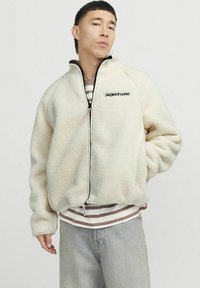 Kreemjas fleece-jakk täis lukuga, kõrge kaelusega ja must logo rinnal. Seljas triibulise särgi ja heleda teksapüksiga.