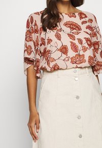 Blouse rose légère à motif floral avec des manches amples, associée à une jupe crème taille haute ornée de boutons argentés.