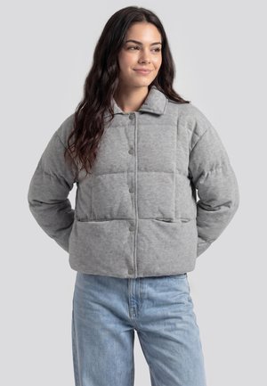 Ung kvinde med langt mørkt hår iført en lysegrå quiltet jakke og lyseblå jeans, kigger let til siden med hænderne bag ryggen.