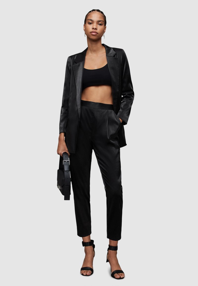 Schwarzer Satinanzug mit einem kurzen Oberteil, taillierter Blazer und schmalen Hosen. Akzentuiert mit schwarzen Riemchenheels und einer strukturierten schwarzen Tasche.