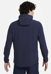 Marinblå huva jacka med ett mjukt, vattentåligt tyg. Har långa ärmar, en avslappnad passform och en rundad fåll. Inga synliga beslag eller mönster.