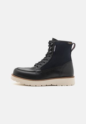 Black leather and textile ankle boot featuring a round toe, lace-up design, and a contrasting cream rubber sole. Includes branding accents.