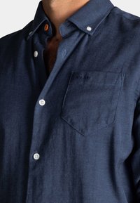 Marineblauw visgraat overhemd met button-down kraag, witte knopen en een borstzak met geborduurd logo. De stof voelt zacht en glad aan.