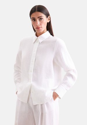 Vrouw met steil donker haar draagt een los wit overhemd met knopen en bijpassende witte broek, staat met handen in de zakken tegen een effen achtergrond.