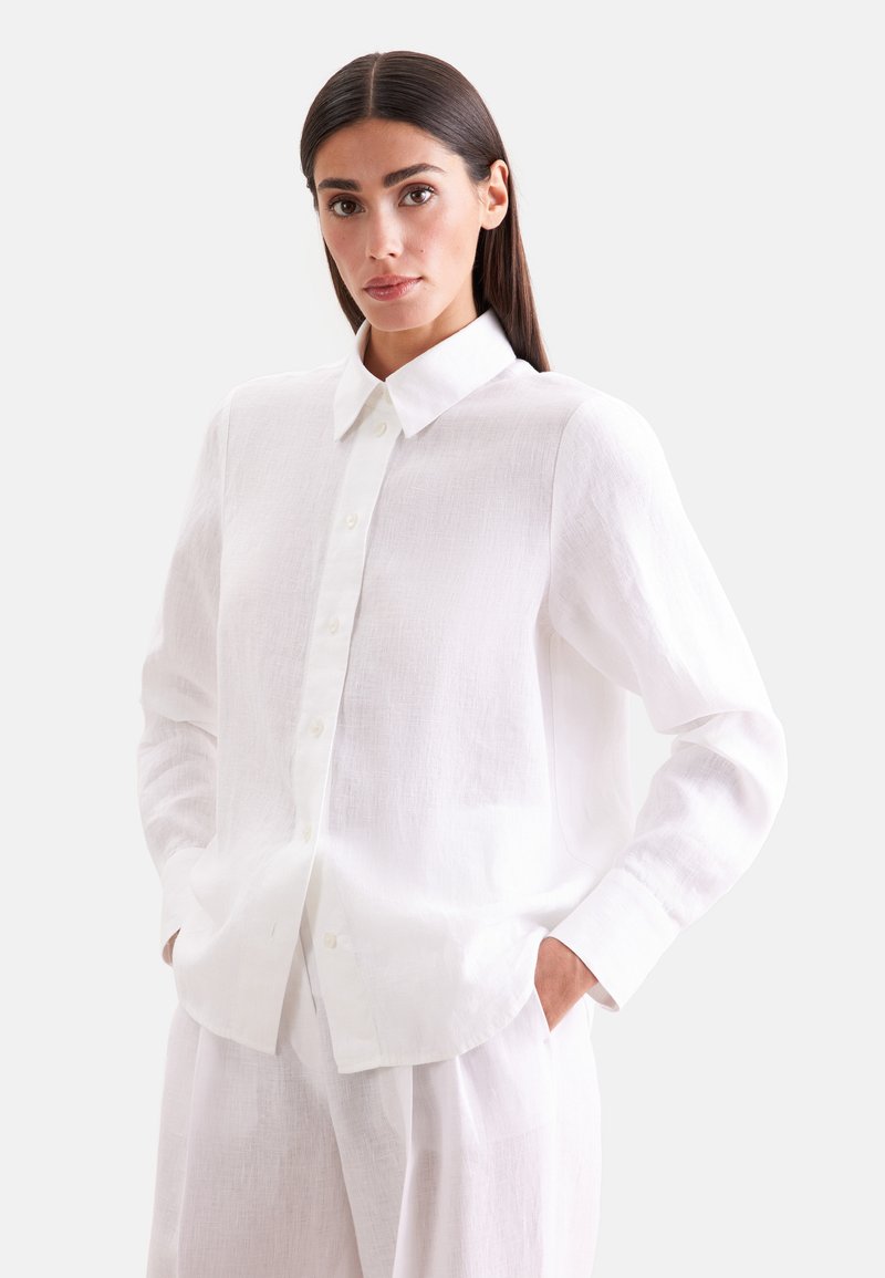 Femme aux cheveux raides et foncés portant une chemise blanche ample à boutons et un pantalon blanc assorti, debout les mains dans les poches devant un fond uni.