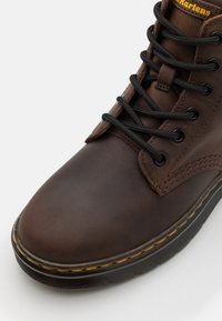 Botte en cuir marron avec des lacets noirs, une couture jaune autour de la semelle et un col rembourré au niveau de la cheville sur fond blanc.