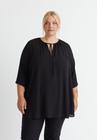 Schwarze Bluse mit einem Schlitz-Ausschnitt, Bindegürtel und lockerer, fließender Silhouette. Hergestellt aus einem glatten, leichten Stoff mit kurzen Ärmeln.
