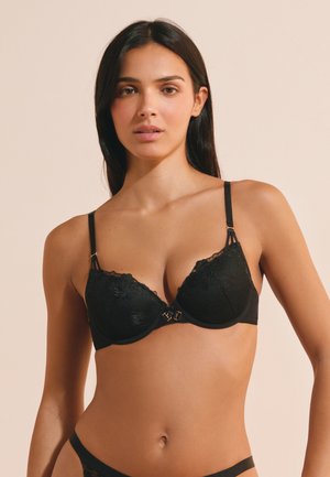 Mujer con cabello oscuro y largo que lleva un sujetador de encaje negro y ropa interior negra a juego sobre un fondo beige liso.