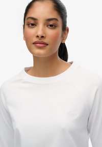 Witte, geborstelde katoenen sweatshirt met een brede halslijn en raglan mouwen. Heeft subtiele branding geborduurd op de borst. Gladde textuur.