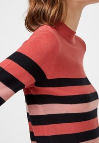 Femme portant un pull col roulé ajusté à manches longues avec des rayures horizontales noires et roses sur un fond rouge.