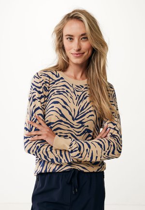 Beige sweater met een marineblauwe tijgerprint, ronde hals en lange mouwen. Zachte, gebreide textuur. Gecombineerd met donkere broek met elastische trekkingkoord.
