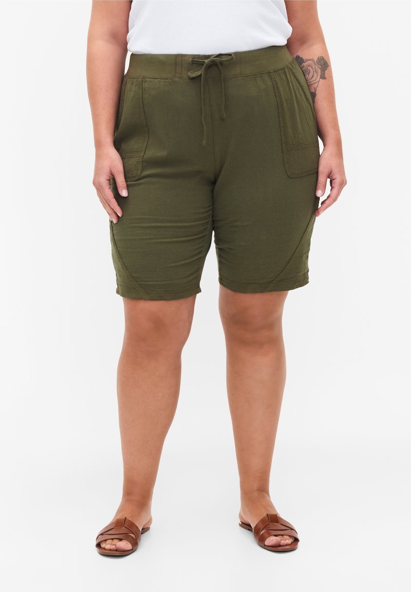 Personne portant un short vert olive à cordon avec poches à l'avant, un haut blanc et des sandales marron, debout sur un fond blanc uni.