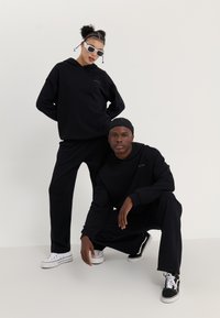 Ensemble de sweat à capuche et de pantalons assortis noirs, fabriqué en tissu doux, avec un design minimaliste sans motifs. Porté avec des baskets décontractées.