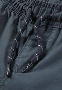 Gros plan sur une ceinture élastique noire avec un cordon noir orné de bandes réfléchissantes blanches sur un pantalon en tissu sombre.
