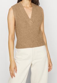 Gebreide mouwloze trui met V-hals in lichtbruine kleur, voorzien van een geribbelde textuur en een aansluitend ontwerp, gecombineerd met crèmekleurige broek.
