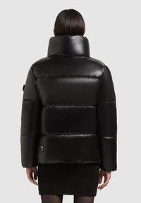 Melnā pūkainā jaka ar augstu apkakli, spīguļojošs materiāls, izšūts dizains un sānu logotipa detaļa. Zem tās redzama tekstūras ribiņa džemperis.