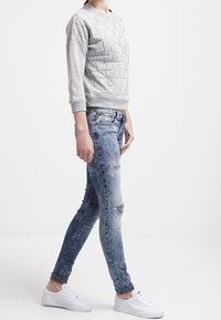 Sudadera gris texturizada con cuello redondo, combinada con jeans ajustados azules desgastados que presentan desgastes, y zapatillas blancas.