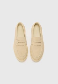 Beige Wildlederslipper mit abgerundeter Spitze, flachem Absatz und einem dekorativen Band über dem Fußrücken. Glatte Textur mit sichtbaren Nähten.