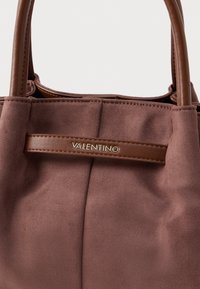 Bolsa em camurça castanha com alças de couro, apresentando uma placa de logotipo dourada e uma textura suave. Costura visível e pregas subtis no corpo.