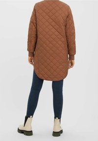 Manteau long matelassé marron avec un ourlet arrondi, porté sur un jean skinny bleu foncé, associé à des bottines beiges avec des semelles noires.