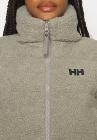 Femme portant une veste polaire gris clair à fermeture éclair avec un col montant et un logo noir "HH" sur le côté gauche de la poitrine.