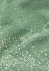 Vihreä kangas, jossa on teksturoitu pinta ja hajallaan oleva vaalean kullan sävyinen kuvio. Mukana teksturoitu vihreä etiketti, jossa lukee "ombre wear."