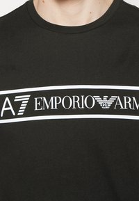 Svart bomullströja med en djärv vit grafik som inkluderar "A7 EMPORIO" och en örnlogotyp, standard rund halsdesign.