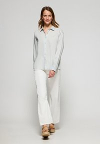 Ljusblå skjorta med långa ärmar, kombinerad med vita vida byxor. En mjuk textur och enkel design har en subtil logotyp.