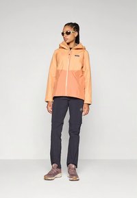 Water-resistant orange and coral jacket with a hood, paired with dark pants and brown hiking shoes. Features visible zippers and logo.