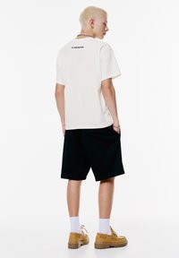 Lys beige t-shirt med et sort "Honda" logo, parret med sorte shorts og gulbrune sko, der står foran en hvid baggrund.