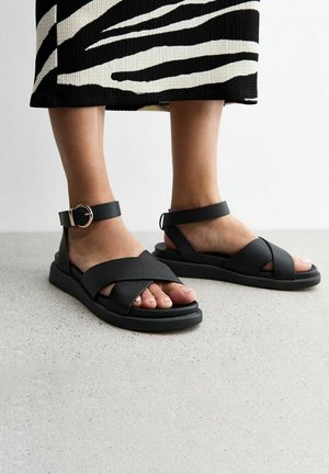 Fødder iført sorte lædersandaler med ankelremme og krydsremme over tæerne, kombineret med et sort- og hvidt zebra-print skørt.