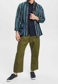 Man in groene wijde broek, marineblauwe sneakers, een gestreept overhemd over een marineblauw shirt, en met een kleine blauwe schoudertas.