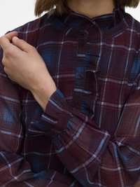 Persoon draagt een bordeauxrood en blauw geruit overhemd met knopen en een ruchesdetail bij de kraag, houdt met de rechterhand de linkerarm vast.