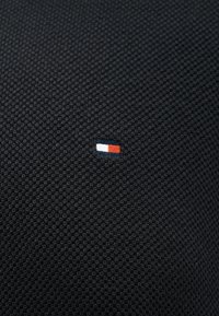 Μικρό ορθογώνιο λογότυπο με οριζόντια μπλοκ σε μπλε σκούρο, λευκό και κόκκινο, κεντημένο σε σκούρο υφασμάτινο φόντο με υφή.