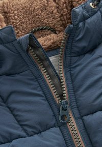 Marineblaue gesteppte Jacke mit einem braunen, fleecegefütterten Innenfutter, einem kontrastierenden Reißverschluss in Bronze und einem strukturierten Stoff mit glatter Oberfläche.