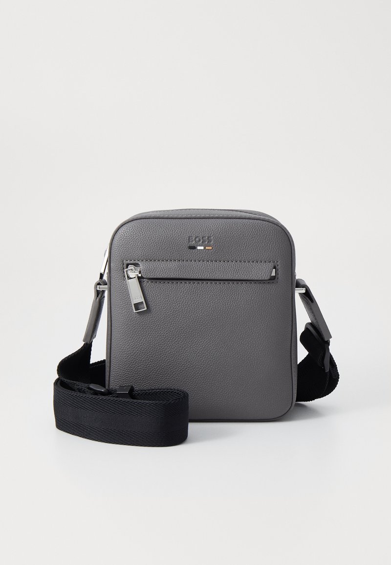 Γκρι τσάντα χιαστί από ανάγλυφη δερμάτινη υφή με επάνω φερμουάρ, μπροστινή τσέπη και μαύρη ρυθμιζόμενη λουρί. Ορατές λεπτομέρειες ασημένιου υλικού.