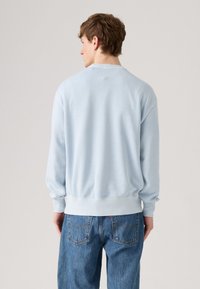 Jonge man die weggedraaid staat, gekleed in een lichtblauwe sweater met lange mouwen en blauwe spijkerbroeken, tegen een effen achtergrond.
