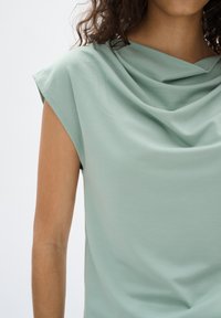 Top drappeggiato verde chiaro con scollo a cowl e maniche corte. Il tessuto appare liscio, leggermente strutturato e morbido al tatto.