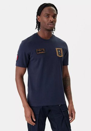 Uomo con capelli intrecciati che indossa una maglietta blu scuro con toppe con la scritta "1 Leader" e uno stemma, abbinata a pantaloni cargo scuri, in piedi contro uno sfondo bianco.