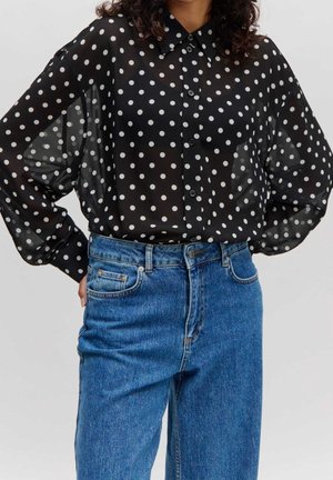 Vrouw draagt een zwarte doorschijnende blouse met witte stippen en knopen, ingestopt in een blauwe high-waisted spijkerbroek.