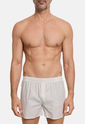 Bar overkrop mand iført løse boksershorts i lysegrå og hvid striber med knapper foran og elastik i taljen, stående mod en hvid baggrund.