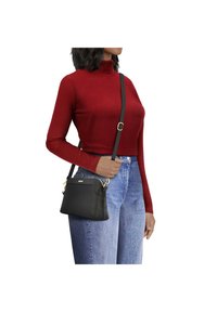 Schwarze Lederüberraschungstasche mit goldenen Beschl ägen, rechteckiger Form, glatter Textur und verstellbarem Gurt, getragen mit einem roten gerippten Pullover.