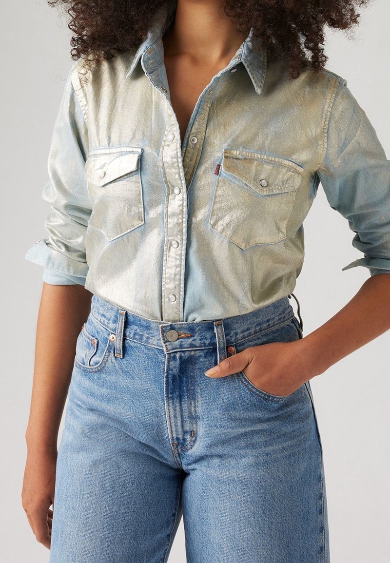 Femme portant une chemise en denim bleu clair à reflets métalliques et un jean bleu taille haute, une main dans la poche.