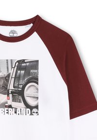 Biała koszulka z maroonowymi rękawami raglanowymi, z czarno-białą grafiką vintage pojazdu oraz napisem "Timberland" na przodzie.